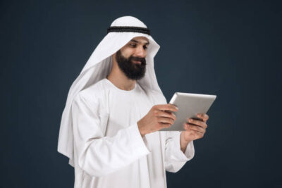 Half-length portrait of arabian saudi businessman on dark blue studio background. Young male model using tablet or gadget. Concept of business, finance, facial expression, human emotions, technologies.